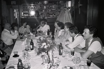 Black-and-white group dinner in a dimly lit, rustic indoor setting—likely a 1970s European gathering. Long table laden with w...
