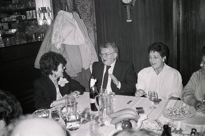 Black-and-white wedding reception scene featuring three central guests: a man in a suit laughing, flanked by women in formal ...