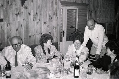 Mid-century indoor gathering at a wooden-paneled dining table, featuring five adults seated/standing around a wine-laden tabl...