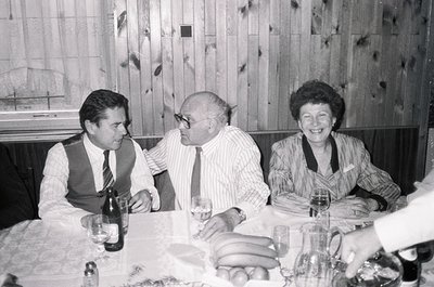 Black-and-white photo of three adults seated at a formal dining table, likely from the **1970s**. Wood-paneled walls and vint...