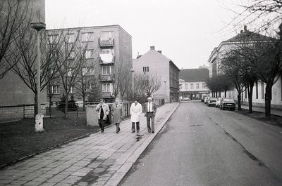 Three men in winter coats walk along a paved sidewalk beside a residential street lined with mid-century concrete apartment b...