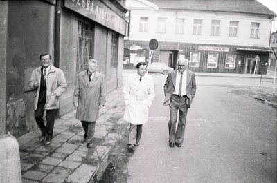 Four individuals in 1960s-era attire—men in coats and ties, one woman in a white coat—walk along a wet urban sidewalk. Soviet...