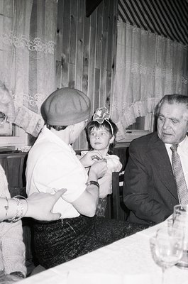 Black-and-white indoor scene from the 1960s–70s featuring a man in a suit adjusting a child’s hat, likely at a formal event. ...