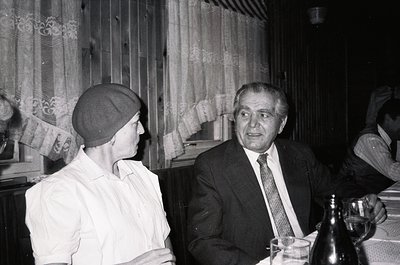 Two individuals in mid-20th-century formal attire converse indoors, likely at a social gathering. The man wears a suit, tie, ...