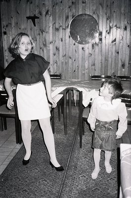 Two children in a wooden-paneled indoor setting, likely a 1960s-70s dining area. The girl in a black collar dress and white s...