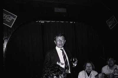 Man in formal attire speaking at a podium under stage lighting, flanked by floral arrangements. Coca-Cola signage visible in ...