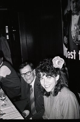 Black-and-white candid shot of two people at an indoor event, likely a 1970s–1980s social gathering. The woman wears a flower...