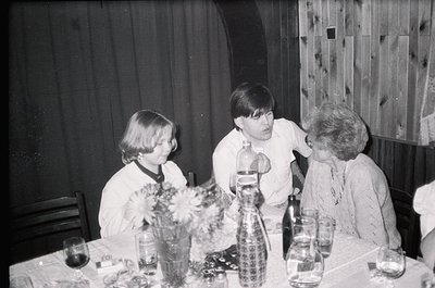 Three individuals seated at a table in a mid-century indoor setting, likely a 1960s-1970s home or gathering. The woman on the...