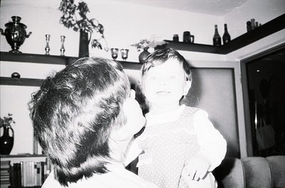 Mid-20th century indoor portrait: an adult and child share a tender moment in a cluttered living room. Shelves display glassw...