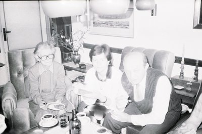 Three adults seated indoors in a mid-century living/dining area, likely 1960s–1970s. Glasses, plates, and a wine bottle sugge...