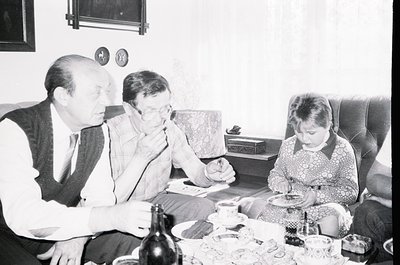 Family gathering in a mid-century living room, featuring three adults and a child seated around a low coffee table. The adult...
