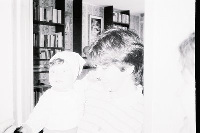 Vintage black-and-white photo of a child in mid-1960s/70s domestic setting, holding a stuffed animal near a bookshelf filled ...