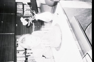 Vintage black-and-white shot of a child sitting on a wooden floor, surrounded by stacked bookshelves. A vase with a single wh...