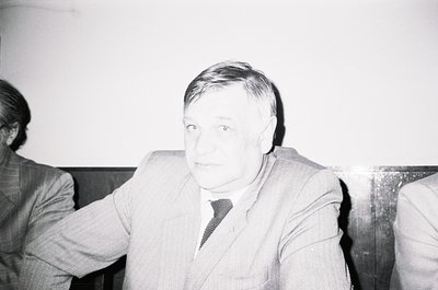 Black-and-white portrait of a man in a formal suit and tie, seated indoors with blurred background. His expression is neutral...
