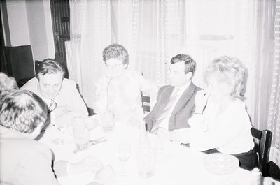 Five adults seated around a table in a dimly lit indoor setting, likely a formal gathering or meeting. Men wear suits, women ...