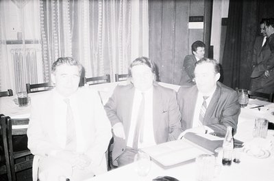 Three men in formal attire—dark suits, ties, and dress shirts—engage in discussion at a rectangular table in an indoor settin...