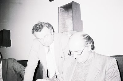 Two men in formal attire examine a geometric sculpture in an indoor gallery. The man on the left wears a suit and glasses, wh...