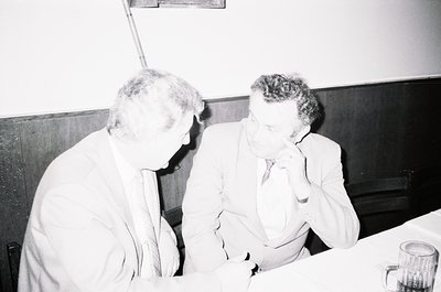 Two men in formal attire—white button-down shirts, ties, and blazers—engage in conversation indoors, likely mid-20th century ...