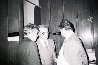 Three men in formal attire—striped suit, white shirt with tie, and light blazer—engage in conversation near a wooden-paneled ...
