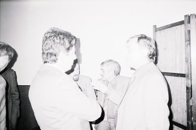 Black-and-white photo of a group of men in formal attire, likely 1960s–1970s, gathered around a projector screen. One man in ...