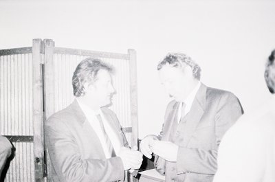 Two men in formal attire exchange a small object at an indoor event, likely mid-1970s–1980s based on clothing styles. The blu...