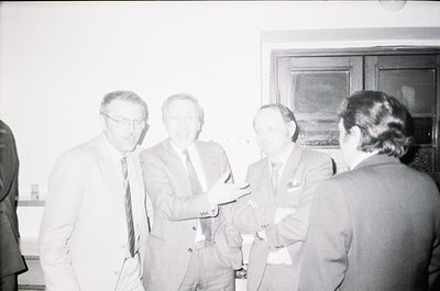 Four men in formal attire—dark suits, ties, and name badges—engage in conversation indoors, likely at a conference or profess...