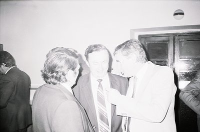 Four men in formal attire—dark suits, ties—engage in discussion indoors, likely a 1970s–1980s conference or office setting. O...