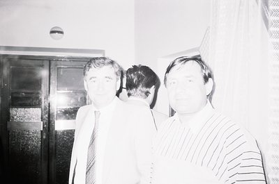 Black-and-white portrait of two men in formal attire, likely 1970s–1980s. The man on the left wears a suit with a patterned t...