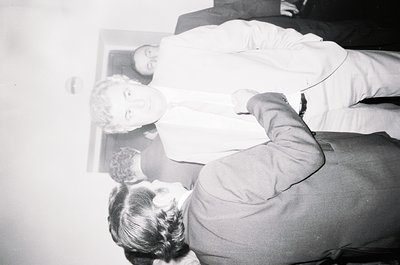 Vintage black-and-white photo of a seated man in formal attire, adjusting a woman’s hair in a mirror. Reflective surface reve...