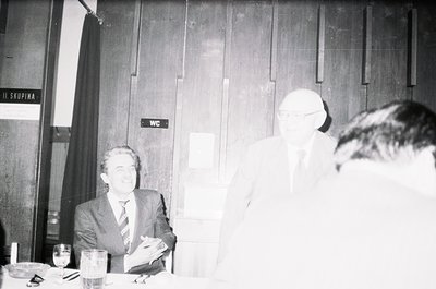 Black-and-white photo of three men in a formal indoor setting, likely a 1970s–1980s conference or meeting. Signage in Cyrilli...