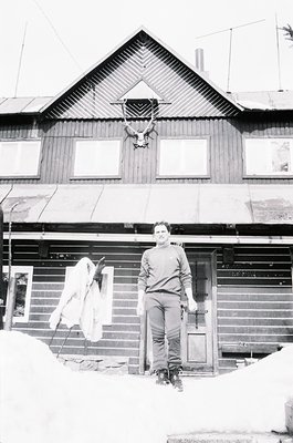 Classic black-and-white photo of a wooden log cabin in snowy terrain. Man in winter gear stands at entrance, holding a rifle....