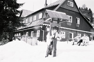 Two men in winter attire pose near a wooden alpine lodge with signage in Cyrillic. Snow blankets the ground and rooftops. One...
