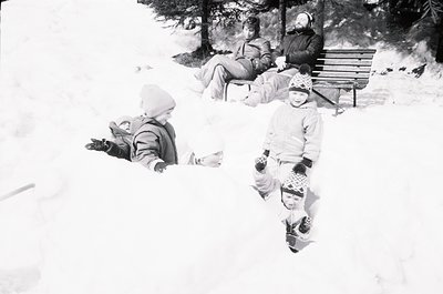 Mid-20th century black-and-white snapshot of winter family fun in alpine setting. Three children build snow structures while ...