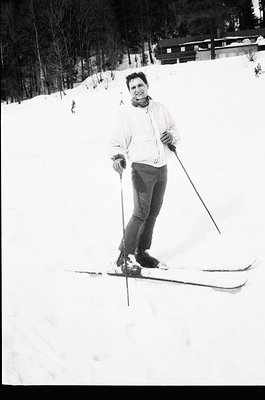 Black-and-white ski portrait of a person in mid-stance on groomed slopes, holding classic skis and poles. Forest and wooden s...