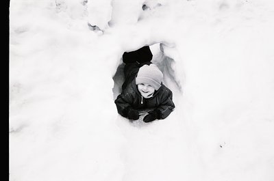 Child buried in snowdrift, smiling up from a dug-out hole. Black-and-white winter scene evoking mid-20th century play. Warm, ...