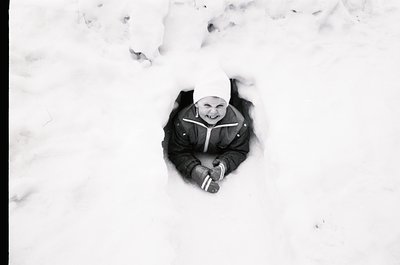 Child buried in snow, smiling up from a shallow hole, wearing a knit hat and winter coat. Black-and-white winter scene evokin...