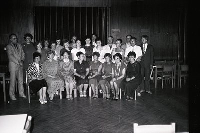 Group portrait in a dimly lit indoor venue, likely a community hall or cultural center, featuring 20+ individuals in 1970s Ea...