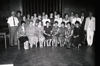 Group portrait from mid-20th century (likely 1950s–1960s) in a formal indoor setting. 18 seated individuals—men in suits, wom...
