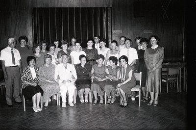 Group portrait of 20+ individuals in a dimly lit indoor setting, likely a community hall or theater. Seated on wooden chairs ...
