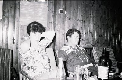 Two individuals seated in a rustic wooden interior, likely mid-20th century. Woman in floral dress leans back, man in striped...