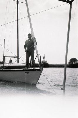 Classic black-and-white sailboat portrait: man in mid-1960s attire stands atop a fiberglass sailboat (PS 517) gripping a haly...