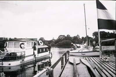 Black-and-white maritime scene featuring a dockside with three boats: a houseboat with "LIVING" signage, a sailboat named "Da...