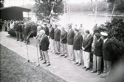 Mid-20th century formal ceremony with men in suits and caps standing in line for a speech or event. Microphone and trees in b...