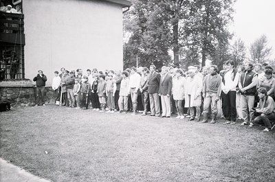 A black-and-white group photo of ~30 individuals standing in a single-file line outdoors, likely mid-20th century. Casual att...