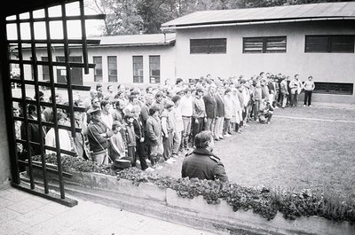 Long queue of people lined up outdoors, separated by a metal fence, near a mid-century institutional building. Uniform attire...