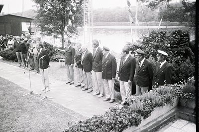 Black-and-white photo of formal outdoor gathering, likely mid-20th century. Group of men in suits, caps, and naval uniforms l...