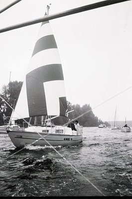 Classic 1970s sailboat "ANIE II" cutting through choppy waters, framed by a dock pole. Mid-century design with striped mainsa...
