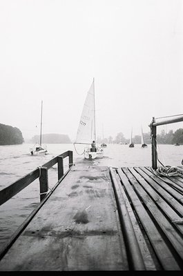 Black-and-white shot of a wooden dock extending into a misty lake, with a single-hander sailboat (number 8277) moored nearby....