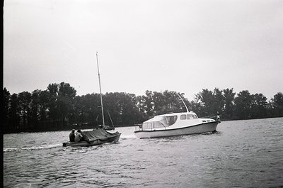 Vintage black-and-white photo of two small boats on calm waters: a motorized cabin cruiser and a simple sailboat with a cover...