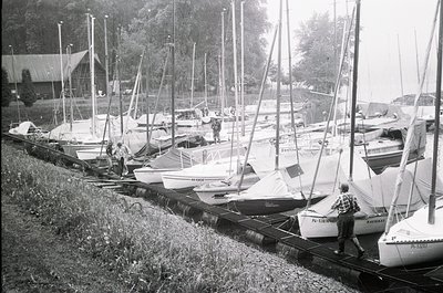 Classic black-and-white marina scene featuring a row of small sailboats and motorboats docked on wooden supports. A man in a ...